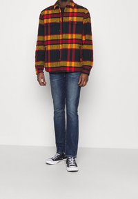 Camicia di flanella a quadri rossa, gialla e nera con chiusura a bottoni, due tasche sul petto, abbinata a jeans blu e sneakers nere.