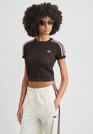 Braunes bauchfreies Oberteil mit strukturiertem Design, weißen Seitenstreifen und gesticktem Logo; kombiniert mit cremefarbenen Hosen mit purpurroten Streifen.