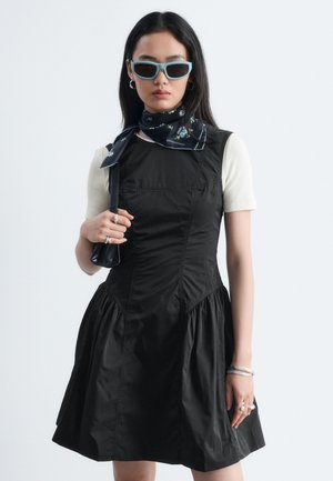 Femme portant une robe noire sans manches sur une chemise blanche à manches courtes, foulard bleu à fleurs autour du cou, lunettes de soleil bleues, tenant un petit sac à main noir.
