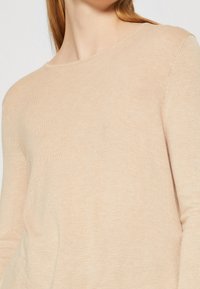 Pull en tricot beige clair avec un col rond et des manches longues. Texture douce avec un fin motif côtelé, indiquant un ajustement confortable.