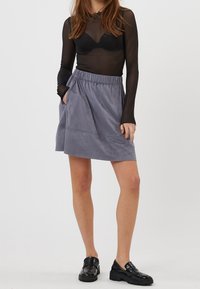 Jupe patineuse grise en tissu doux avec une taille élastique, poches latérales et une silhouette légèrement évasée. Portée avec des chaussures noires et un haut transparent.