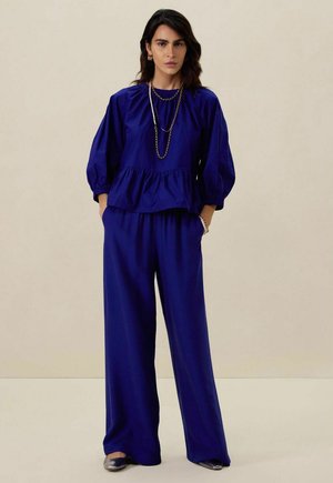 Blouse bleu cobalt avec des manches bouffantes et un détail peplum, associée à un pantalon large assorti ; accents en bijoux en argent simples.