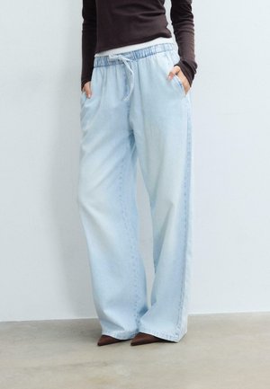 Osoba ubrana w jasnoniebieskie szerokie jeansy z wiązaniem w talii i brązowe, spiczaste buty, ręce w kieszeniach, stoi oparta o gładką ścianę.