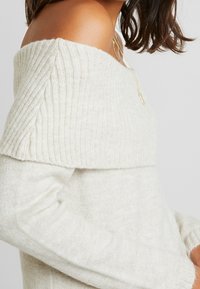 Cremfarbener, schulterfreier Strickpullover mit geripptem Muster, langen Ärmeln und legerem Schnitt. Das leichte Material sorgt für ein weiches, gemütliches Tragegefühl.