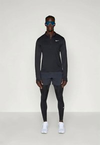 Black long-sleeve athletic top with half-zip and Nike logo, paired with black shorts, tight leggings, and white running shoes. Blue sunglasses.