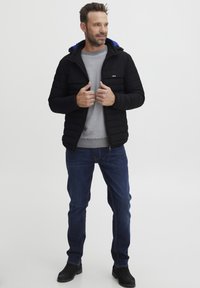 Črna podložena jakna z modrim kapuco, siva rebrasta pulover in modri kavbojke. Oseba nosi črne gležnjarje in stoji pred nevtralno ozadje.