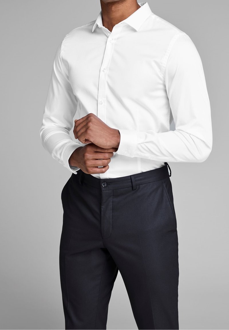 Camicia bianca con colletto e maniche lunghe, dal tessuto liscio, abbinata a pantaloni scuri con frontale piatto e una leggera lucentezza.