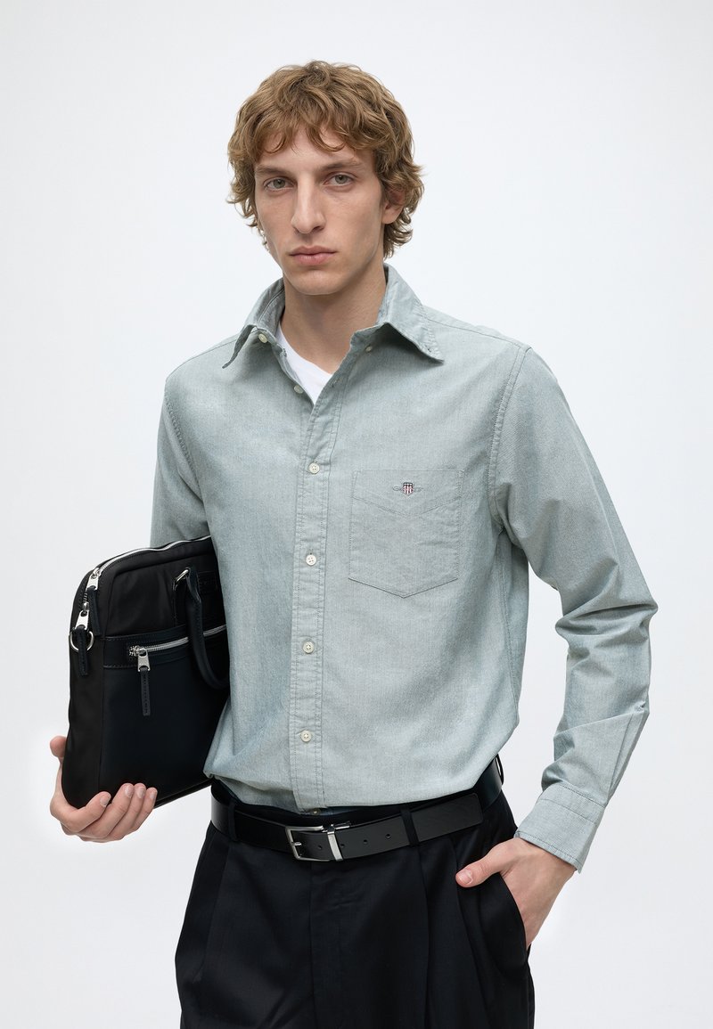 Joven con cabello rizado claro que lleva una camisa gris claro abotonada y pantalones negros, sosteniendo una bolsa negra para portátil sobre un fondo blanco.