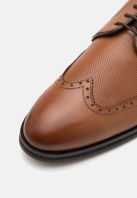 Chaussure habillée en cuir marron avec un design perforé, bout pointu et surpiqûres accentuées. Semelle en caoutchouc noir avec des détails contrastants.