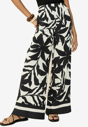 Pantalon noir et blanc à jambes larges avec de grands motifs de feuilles, porté avec des sandales noires et des bracelets à un poignet.