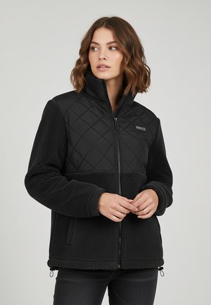 Woman with shoulder-length wavy hair wearing a black quilted and fleece zip-up jacket, looking slightly left, hands clasped in front.