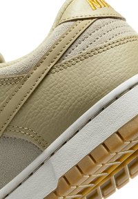 Primer plano de una zapatilla beige que muestra cuero texturizado, parte superior de ante, detalles cosidos y entresuela blanca con suela exterior de goma de goma con patrón.
