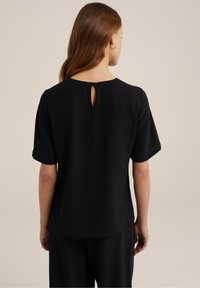 Černý top s krátkým rukávem a texturovaným povrchem, kulatým výstřihem a detaily ve tvaru klíčové dírky na zadní straně. Volný střih zajišťuje pohodlí.