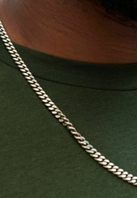 Silberne Kettenhalskette mit ovalen Gliedern, die auf einem dunkelgrünen Shirt liegt. Das Shirt hat einen Rundhalsausschnitt und eine glatte Textur.