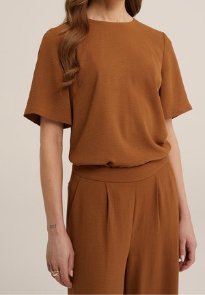 Femme portant un haut marron texturé à manches courtes et un pantalon large plissé taille haute assorti sur un fond neutre.