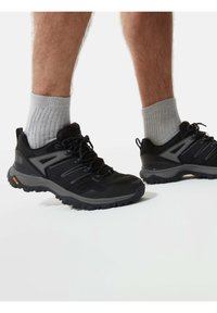 Jambes portant des chaussettes grises et des chaussures de randonnée noires avec des semelles Vibram robustes sur fond blanc.