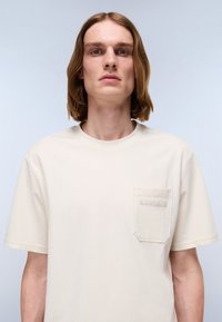 Beige bomull t-skjorte med rund hals, kort erme og en frontlomme med preget logo. Jevn tekstur gjennom hele.
