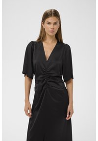 Musta mekko, tehty sileästä kankaasta, jossa syvä V-pääntie, puhvimaiset lyhyet hihat, kiristetty vyötärö ja sivuhalkio.