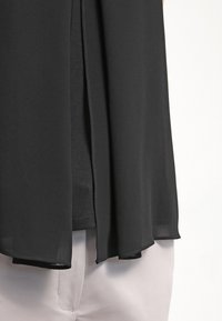 Černá, lehká blůza s volným střihem, s hladkým povrchem a asymetrickým spodním lemem, která lemuje světlé kalhoty.