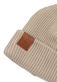 Beżowa czapka dziergana z prążkowaną fakturą, z brązową skórzaną łatą z wytłoczonym wzorem. Materiał jest miękki i elastyczny.