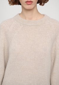 Jeune personne aux cheveux courts et bouclés portant un pull beige doux en tricot avec un col rond côtelé.