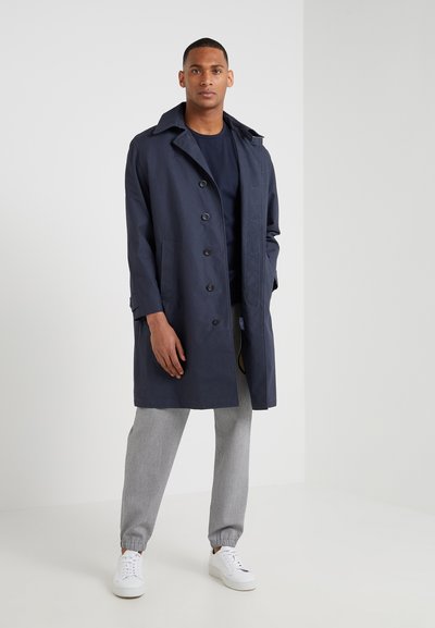 Manteau trench navy avec un col à revers, fermeture à boutons sur le devant et poches latérales, associé à un pantalon tapered gris clair et des baskets blanches.