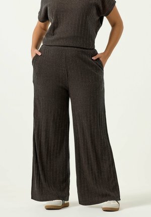 Person iført mørkegrå bredbenede bukser med ribstruktur og matchende kortærmet top, stående med hænderne i lommerne og hvide sneakers.