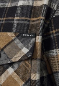 Close-up van bruine, zwarte, grijze en witte geruite stof met een zakflap en een zwart label met de tekst "REPLAY."