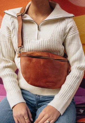 Braune Leder-Umhängetasche mit Reißverschluss, glatter Textur und verstellbarem Riemen, getragen zu einem hellen Strickpullover und Denim.