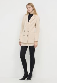 Blazer beige doppiopetto in tessuto testurizzato, dolcevita nero, mini gonna, collant neri e décolleté neri a punta.