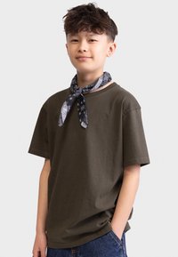 Olivgrünes Baumwoll-T-Shirt mit kurzen Ärmeln, kombiniert mit einem navyfarbenen, gemusterten Schal, der um den Hals gebunden ist. Blaue Jeans aus Denim mit Vordertaschen.