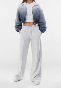 Giacca tagliata, sfumatura di blu a grigio, con polsini elasticizzati; abbinata a pantaloni sportivi grigi chiari a gamba larga, dotati di una vita elastica.