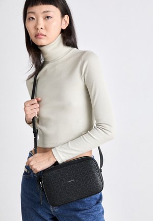 Młoda kobieta ubrana w jasnobeżowy golf i niebieskie dżinsy trzyma czarną torebkę Calvin Klein na ramię z wzorem logo.