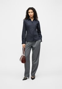 Γυναίκα με σγουρά σκούρα μαλλιά, φοράει μπλε πουκάμισο με κουμπιά, γκρι παντελόνι, μαύρα τακούνια και κρατά καφέ τσάντα, στέκεται μπροστά σε λευκό φόντο.