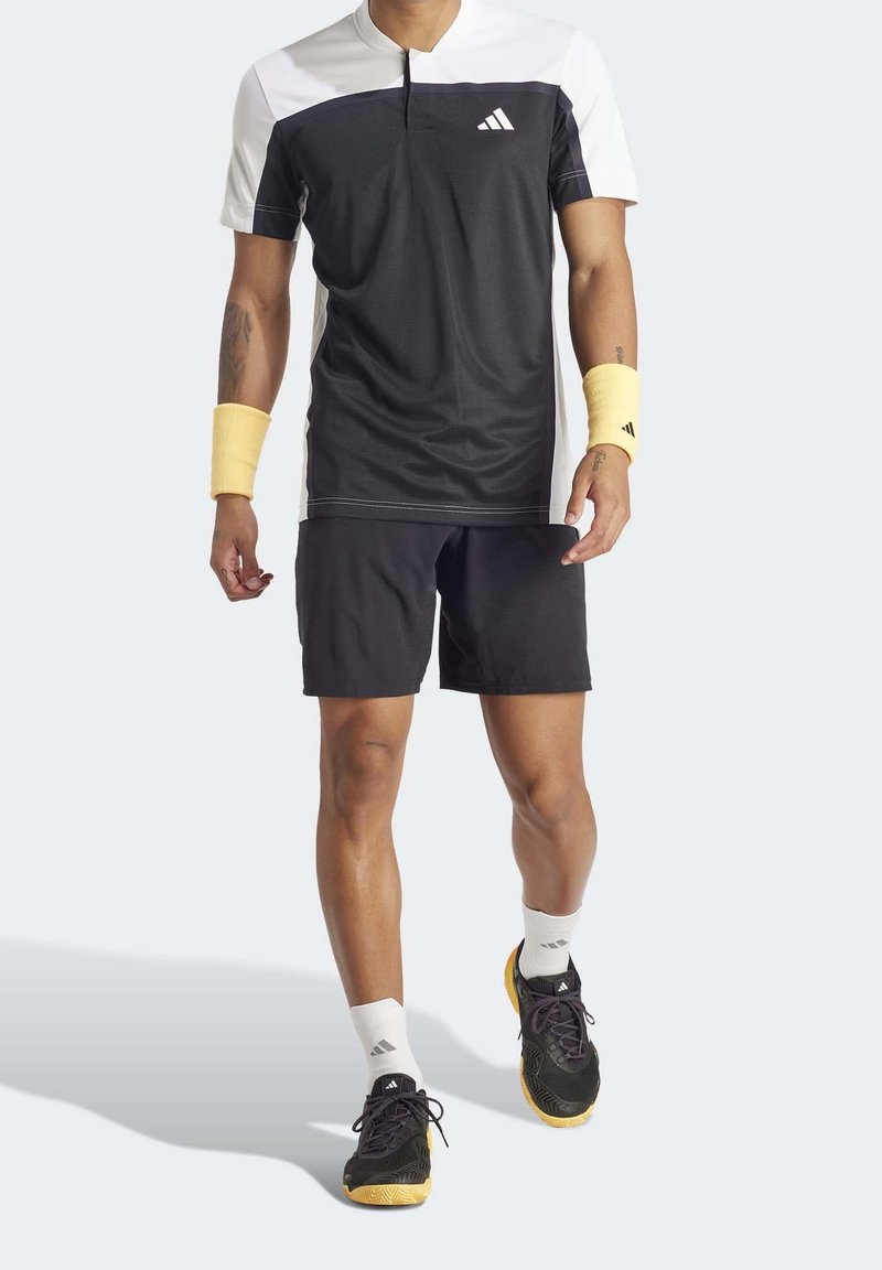 Homme marchant portant un maillot de sport noir et blanc, un short noir, des bracelets jaunes, des chaussettes blanches et des baskets noires et jaunes.