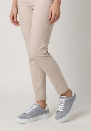 Osoba w beżowych dopasowanych spodniach i jasnoszarych zamszowych trampkach z białymi sznurowadłami, stojąca na jednolitym tle.