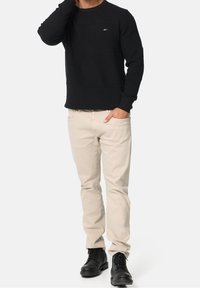 Mann in schwarz strukturiertem Pullover, beigen Hosen und schwarzen Schnürstiefeln, steht mit einer Hand in der Tasche und der anderen Hand nah am Gesicht.
