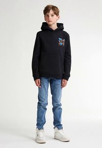 Felpa nera con cappuccio e tasca frontale, con un motivo geometrico multicolore nella parte superiore sinistra. Indossata con jeans azzurri e sneaker bianche.