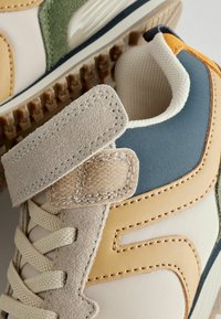 Sneakers med en blanding af beige, grønne og blå materialer. Har tekstureret ruskind, glat læder og en Velcro-strop. Synlige syede detaljer.