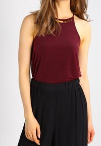 Burgundowa, bezrękawnikowa bluzka z falistym dekoltem i metalowym akcentem, w połączeniu z czarnymi spodniami o wysokim stanie z elastycznym pasem.