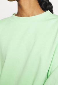 T-shirt in cotone oversize verde menta con scollo rotondo, presenta una texture liscia e una vestibilità rilassata, lasciando scoperta parte della spalla e del collo.