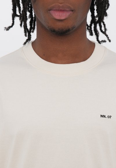 Camiseta de algodón beige con cuello redondo y mangas cortas, que presenta un pequeño logo negro "NN. 07" en el pecho izquierdo. Textura suave.