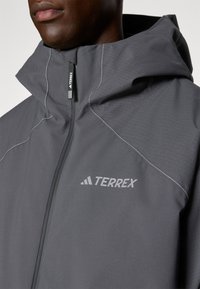 Siva vodootporna jakna s visokim ovratnikom, s reflektirajućim šavovima, patentnim zatvaračem i Terrex logotipom sprijeda.