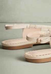 Sandales beiges à semelle plate, avec des fines brides, un nœud texturé en accent, et un bord festonné sur une semelle rembourrée.