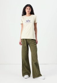 T-shirt en coton beige avec logo "ALPHA INDUSTRIES" violet, associé à un pantalon cargo olive ample avec poches latérales et coupe décontractée.