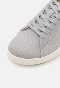 Gray canvas sneaker with textured upper, contrasting light gray laces, padded collar, and cream rubber sole. Subtle stitching detail.