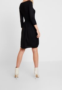 Robe tricotée noire avec corsage côtelé, taille ajustée, jupe fluide, manches trois-quarts et texture lisse ; associée à des bottines beige.