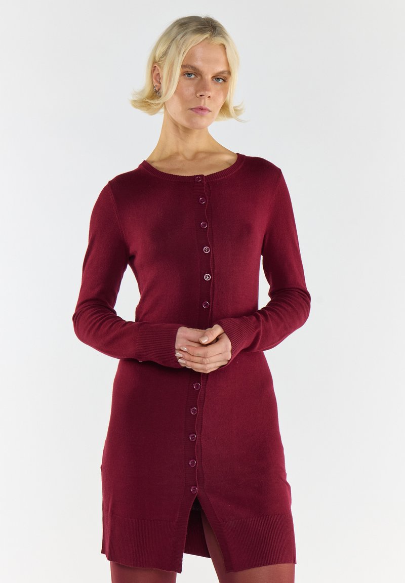 Blondynka w dopasowanej bordowej sukience kardiganowej z guzikami stoi z rękami złożonymi na tle gładkiej białej ściany.