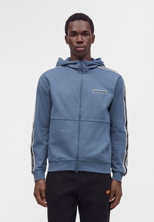 Jeune homme face à l'objectif portant un sweat à capuche bleu zippé avec des bandes de marque sur les manches et un pantalon noir sur un fond uni.