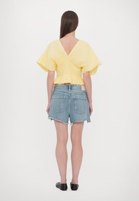 Gelbes Puffärmeloberteil mit V-Ausschnitt, gerafftem Bund und Rüschensaum, kombiniert mit hoch taillierten blauen Jeansshorts und schwarzen Ballerinas.